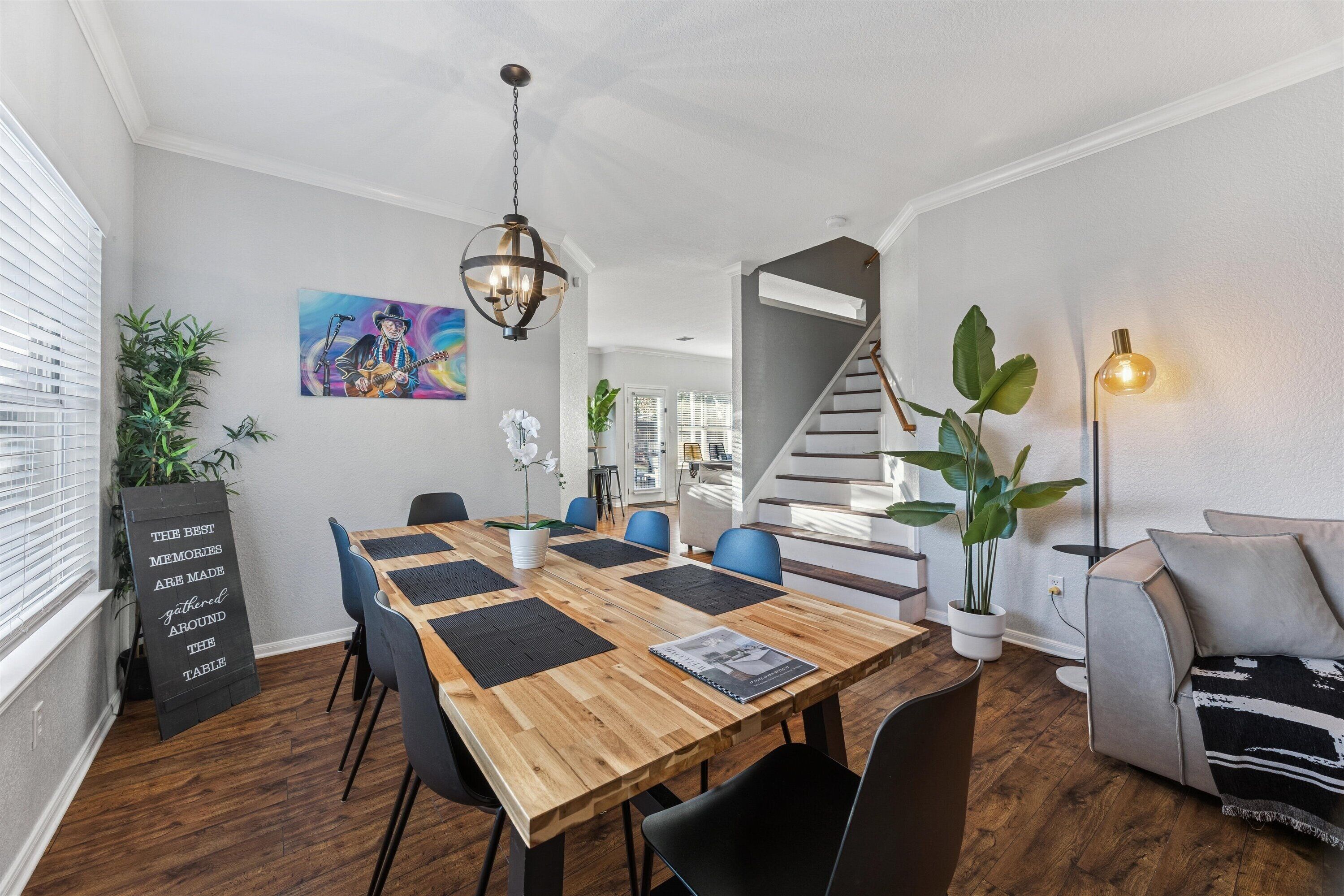 Uma área de refeições com mesa de madeira, cadeiras em preto e branco, um lustre pendurado e uma escada ao fundo.