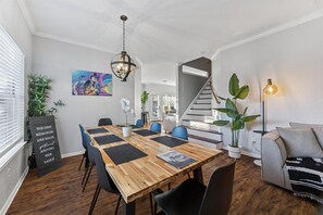 Uma área de refeições com mesa de madeira, cadeiras em preto e branco, um lustre pendurado e uma escada ao fundo.