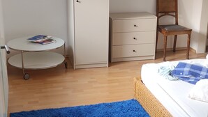 Una habitación con alfombra azul, un armario blanco, una pequeña mesa redonda con libros y una silla de mimbre.
