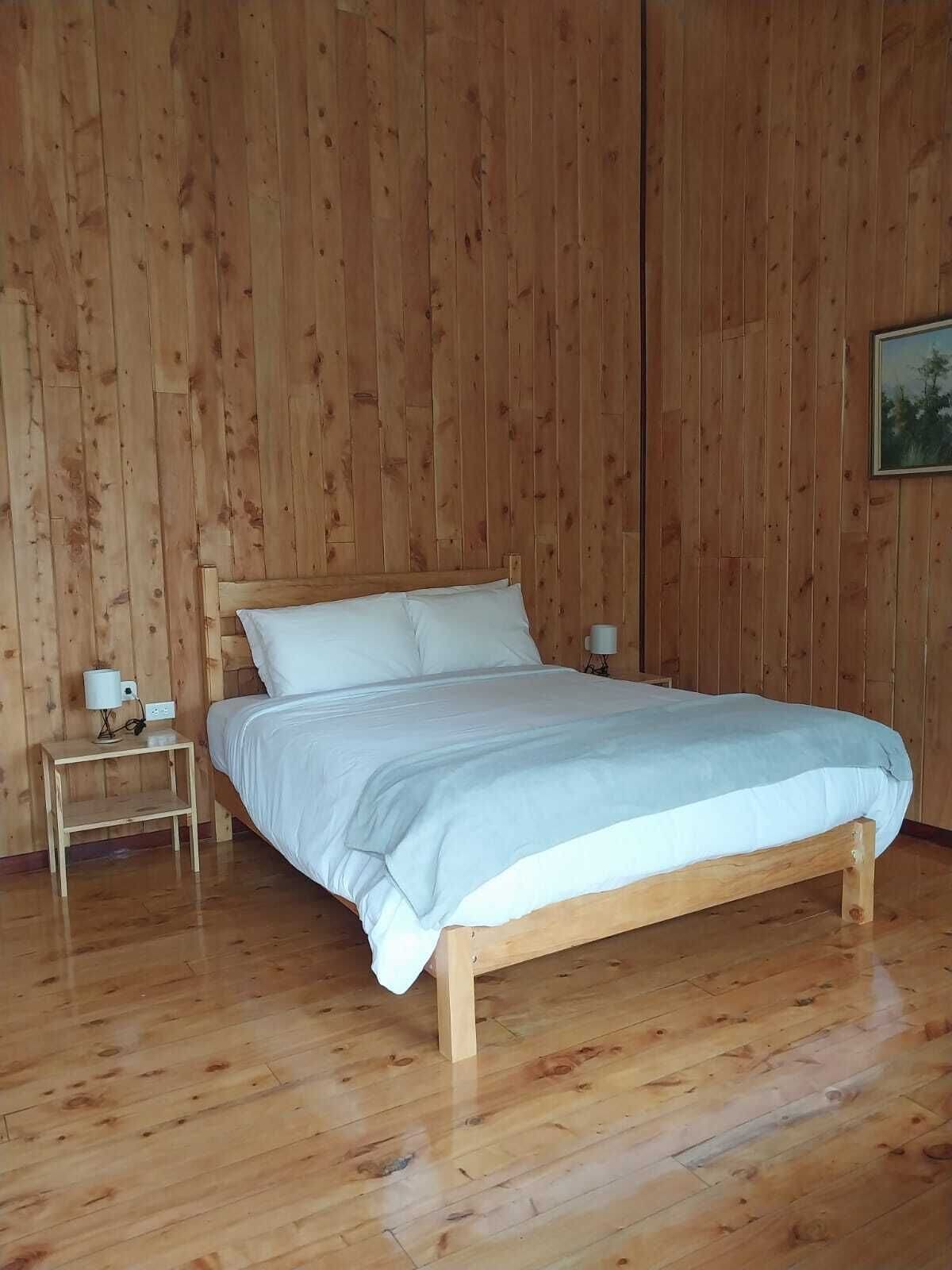 Una habitación con paneles de madera, una cama, una mesita de noche con lámpara y un cuadro en la pared.