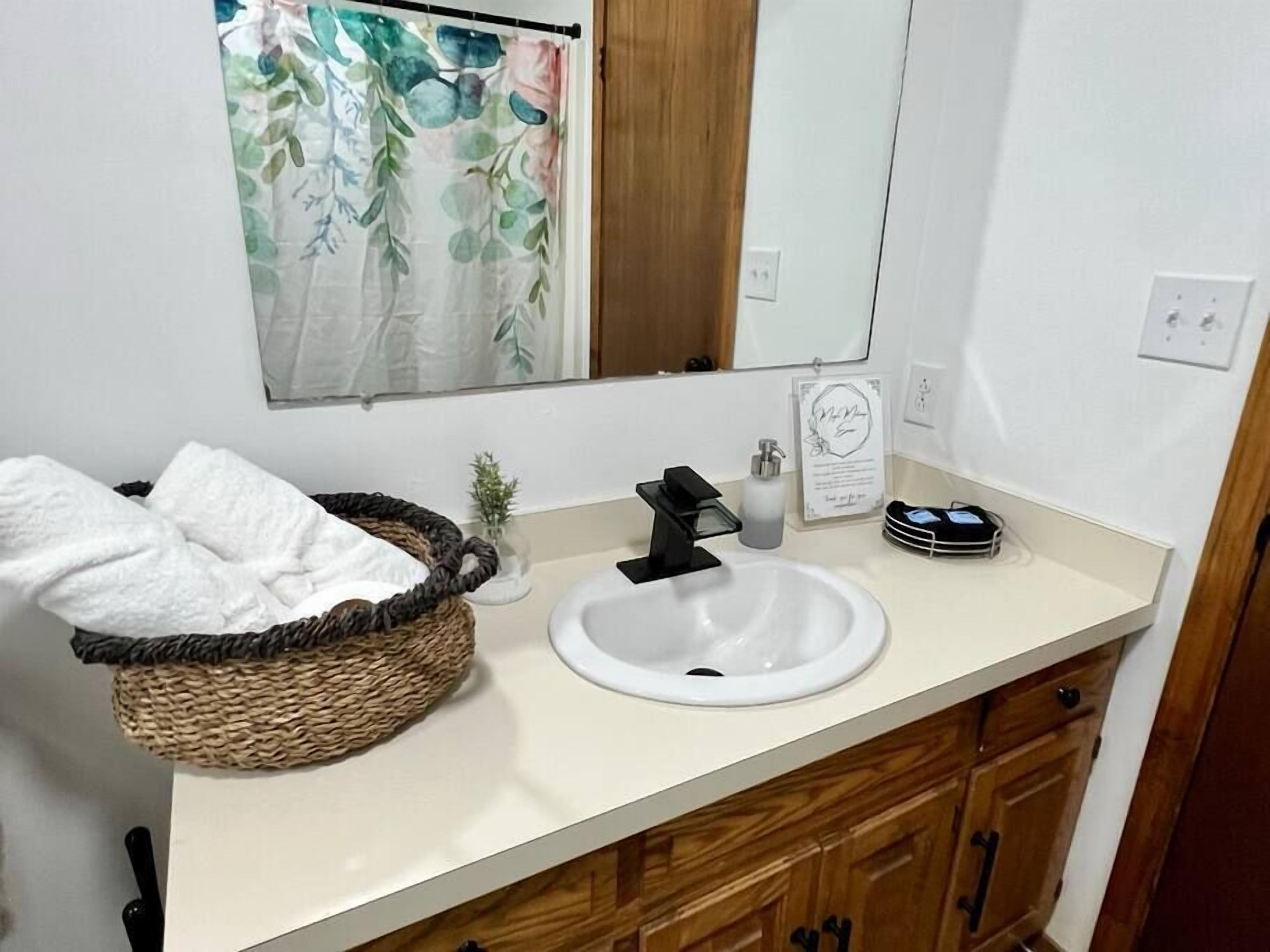 A bathroom with a white sink, a black faucet, a wicker basket with a white towel, a small potted plant, and a mirror above the sink.