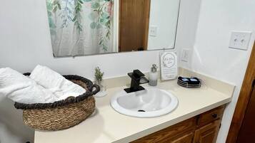 A bathroom with a white sink, a black faucet, a wicker basket with a white towel, a small potted plant, and a mirror above the sink.