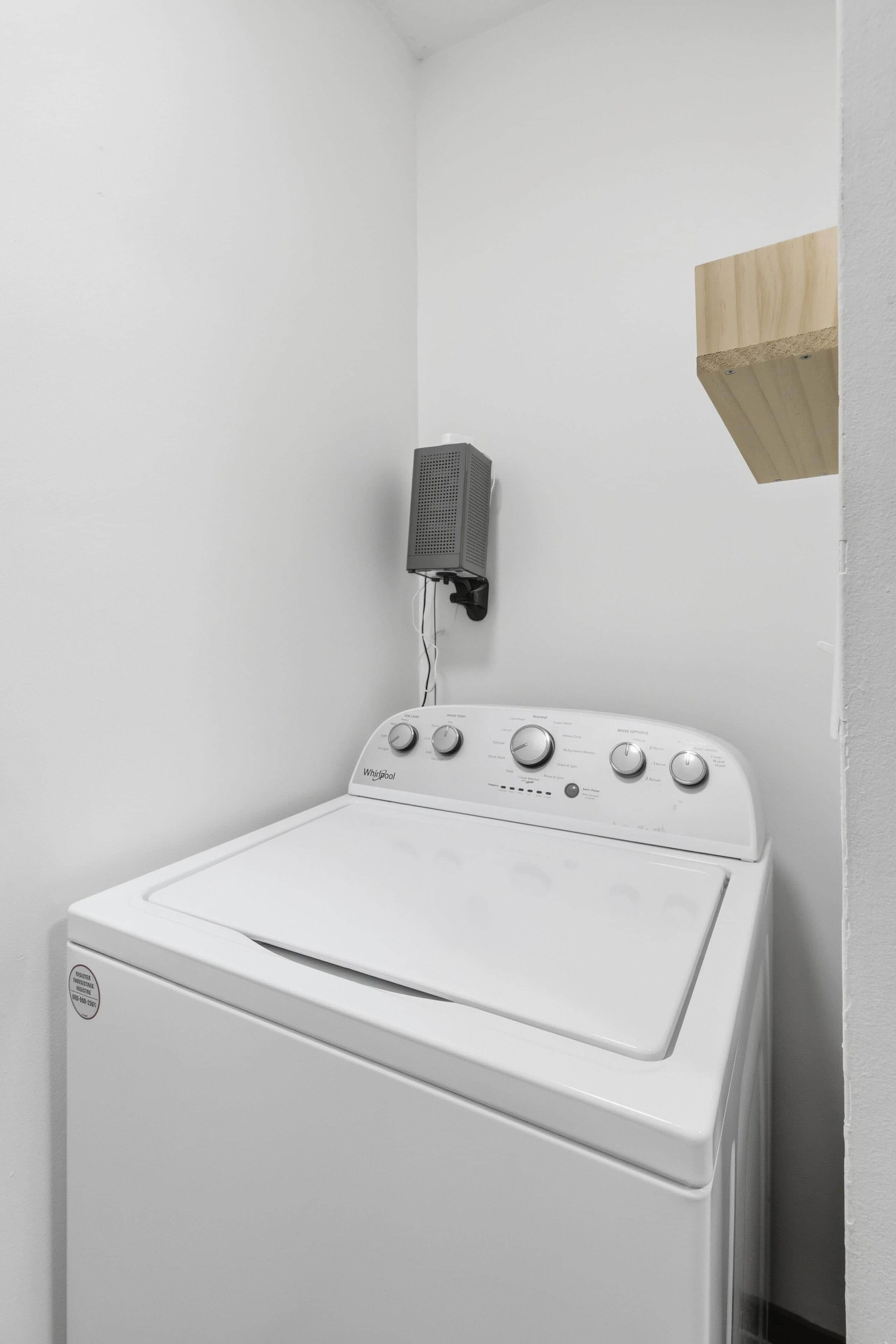 Een witte Whirlpool wasmachine in een hoek van een kamer met witte muren en een houten plank.