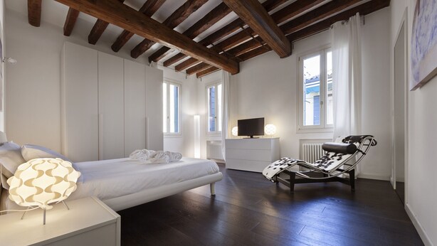 Une chambre à coucher comprenant un lit, une table de chevet, une lampe, une chaise, un téléviseur et des fenêtres.