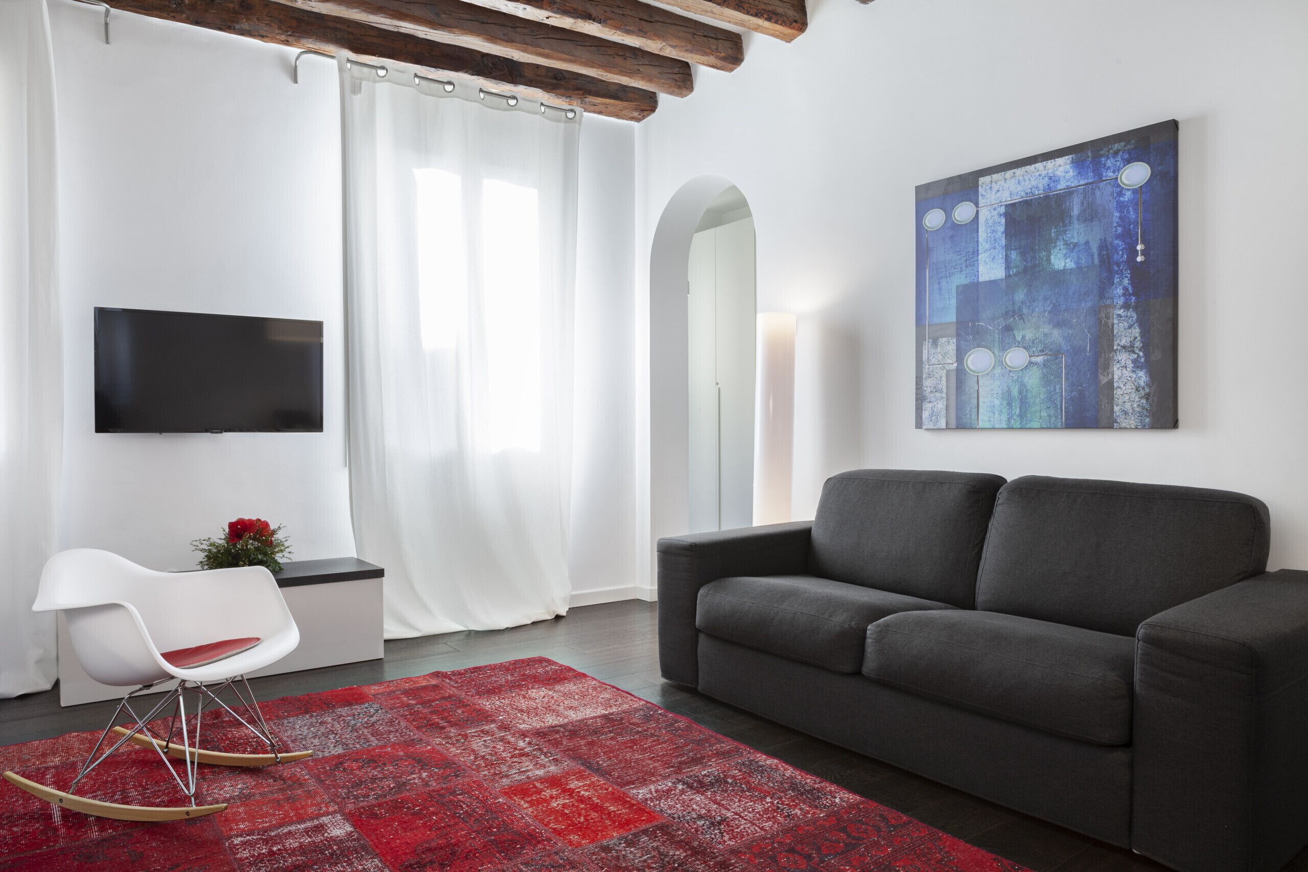 A modern living room with a grey sofa, a white rocking chair, a red rug, a flat-screen TV, and a large abstract painting on the wall.