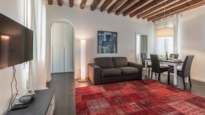 Un salon moderne doté d’un téléviseur à écran plat, d’un canapé gris, d’un tapis à motifs rouges et d’un coin repas avec une table et des chaises.