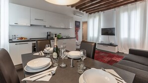 Une cuisine moderne avec des meubles blancs, un coin repas avec une table dressée pour quatre personnes, et un salon avec un téléviseur et un canapé.
