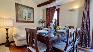 Une salle à manger avec une table en bois dressée pour quatre personnes, entourée de chaises rembourrées. Un tableau est accroché au mur, au-dessus de la table, et une lampe est visible sur la gauche.