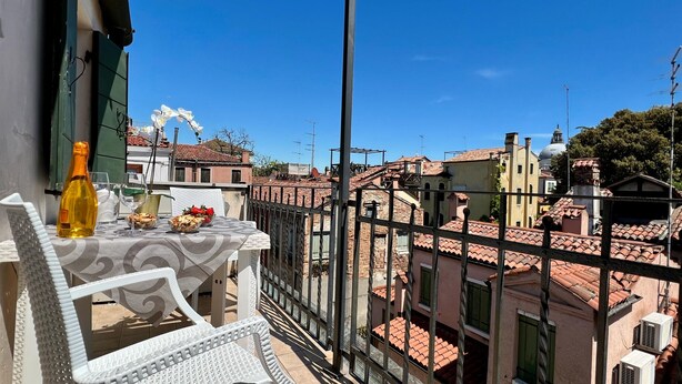 Un balcon avec une table dressée pour deux personnes, une bouteille de vin et un panier de fruits. Le balcon offre une vue sur le paysage urbain, avec ses bâtiments traditionnels et un ciel bleu dégagé.