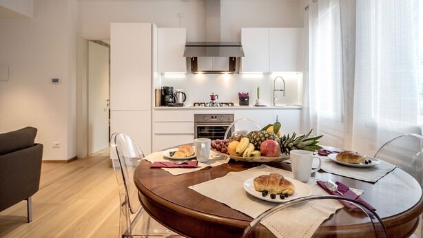Una cuina moderna amb armaris blancs, una campana extractora d'acer inoxidable i un esmorzar a la taula de menjador amb croissants, fruita i una cafetera.