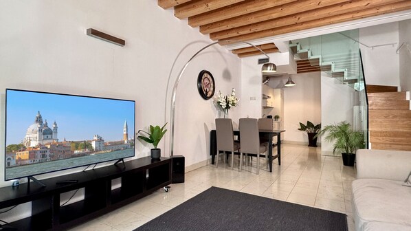 A modern living room with a flat-screen TV, a dining area with chairs and a table, and a staircase leading to an upper level.