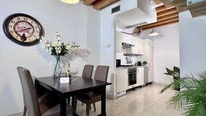 A dining area with a table set for two, a clock on the wall, and a kitchen in the background.