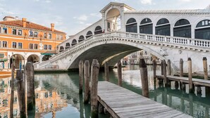 A historic stone bridge with arched walkways over a canal, wooden posts, and a wooden dock extending into the water. - Corner Canal View (300m from Rialto´s Bridge) (Venecia)