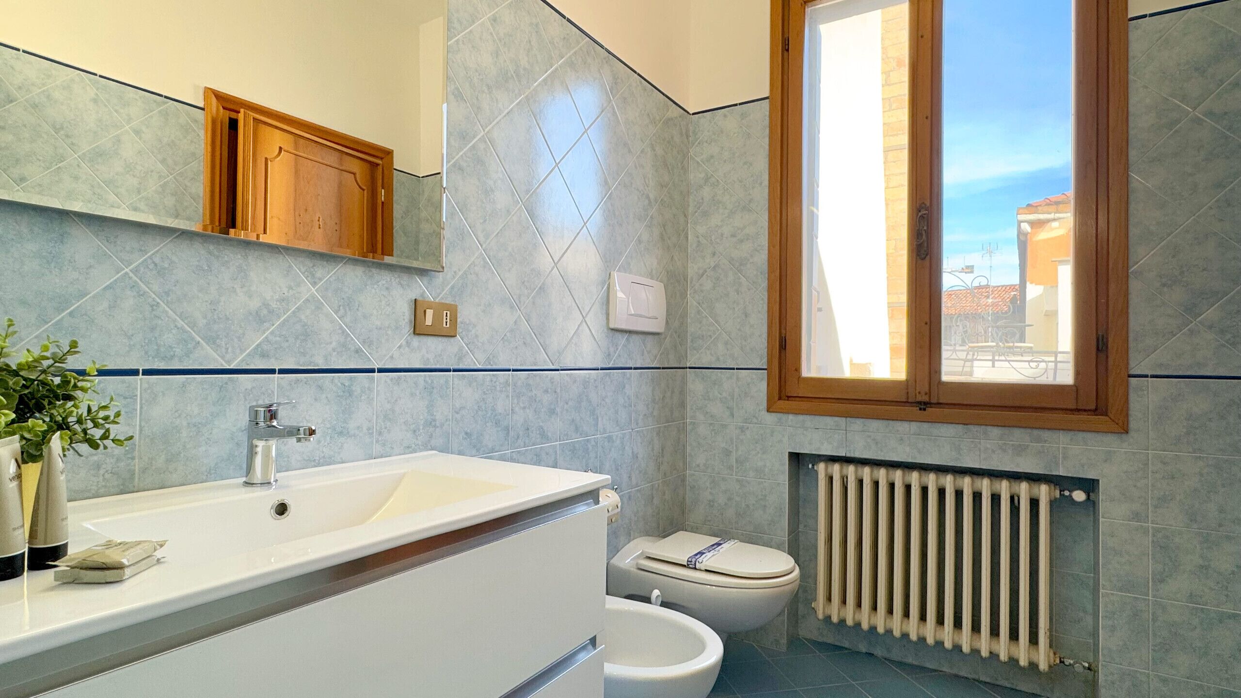 A bathroom with a white sink, a toilet, a radiator, and a window with a view of buildings.