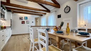 Une cuisine moderne avec des meubles blancs, des poutres en bois et un coin repas agrémenté d’une table en bois mise pour deux personnes.