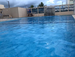 En swimmingpool med ett vattenfall, omgiven av kaklade väggar och ett avskärmat sittområde.