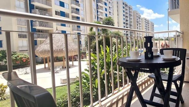 A balcony with a table and chairs, overlooking a pool area with a thatched umbrella and lush greenery. - 207 AKUA RESORT® VACATION APARTMENT (Ricaurte)
