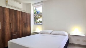 A bedroom with a bed, white bedding, a wooden wardrobe, a window with a view, and bedside lamps. - 207 AKUA RESORT® VACATION APARTMENT (Ricaurte)