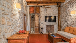 A cozy room with stone walls, a fireplace, a wooden table with a basket of fruit, a sofa, and a flat-screen TV mounted above the fireplace. - Macchiaroli La Gioiosa Castle the magic of a medieval castle in Teggiano (Teggiano)