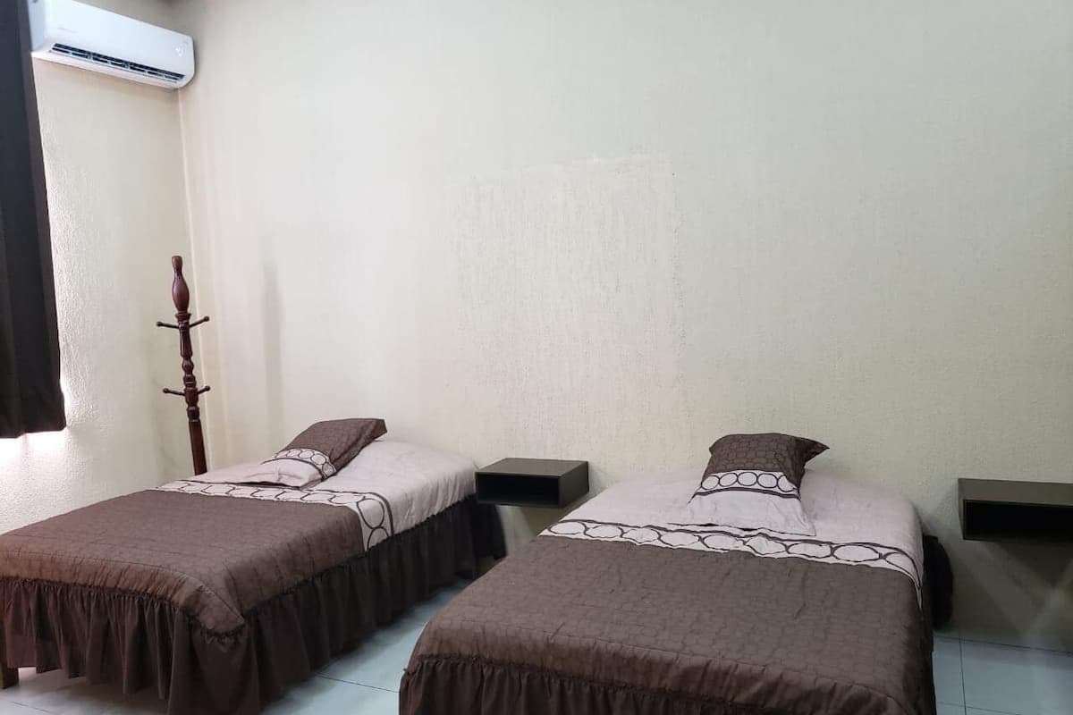 A room with two beds, each with a brown bedspread and a white pillow. There is a wooden coat rack against the wall and a small shelf on the right side.