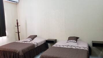 Una habitación con dos camas, cada una con una colcha marrón y una almohada blanca. Se observa un perchero de madera apoyado en la pared y una pequeña estantería a la derecha.