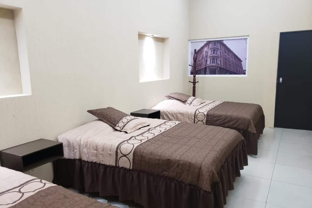 A hotel room with two beds, a nightstand, a lamp, a window with a view of a building, and a door.