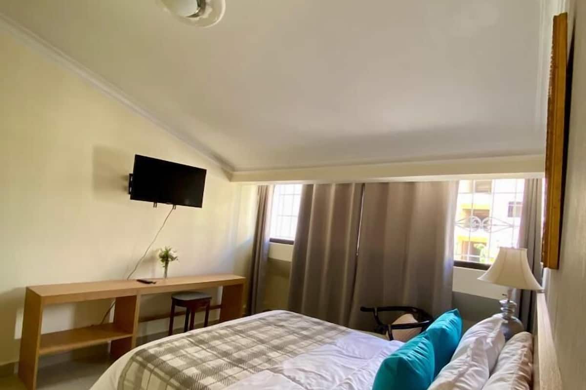 Een slaapkamer met een bed, een bureau, een stoel, een aan de muur bevestigde televisie en een raam met gordijnen.