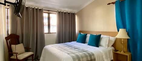 Habitación de hotel con cama, una silla, una mesita de noche, ventilador de techo y televisor montado en la pared.