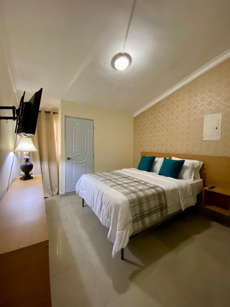 A bedroom with a bed, a nightstand, a lamp, a door, and a wall-mounted television.