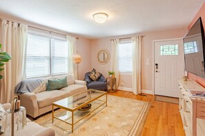 A living room with a beige sofa, a blue armchair, a glass coffee table, and a patterned rug. There is a round mirror on the wall and a television on the right. - Altavista House - Renovated, Charming, & Walkable (Charlottesville)