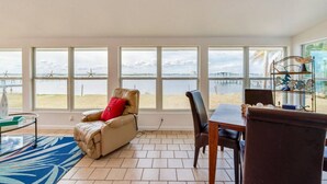 Ein Zimmer mit großem Fenster mit Blick auf Strand und Steg, ein Ledersofa, ein Esstisch und ein Metallregal mit Dekorationen.