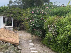 A pathway leading to a white building with a gated entrance, surrounded by lush greenery and flowering plants. - The house of happiness (BAGNOLS EN FORET)