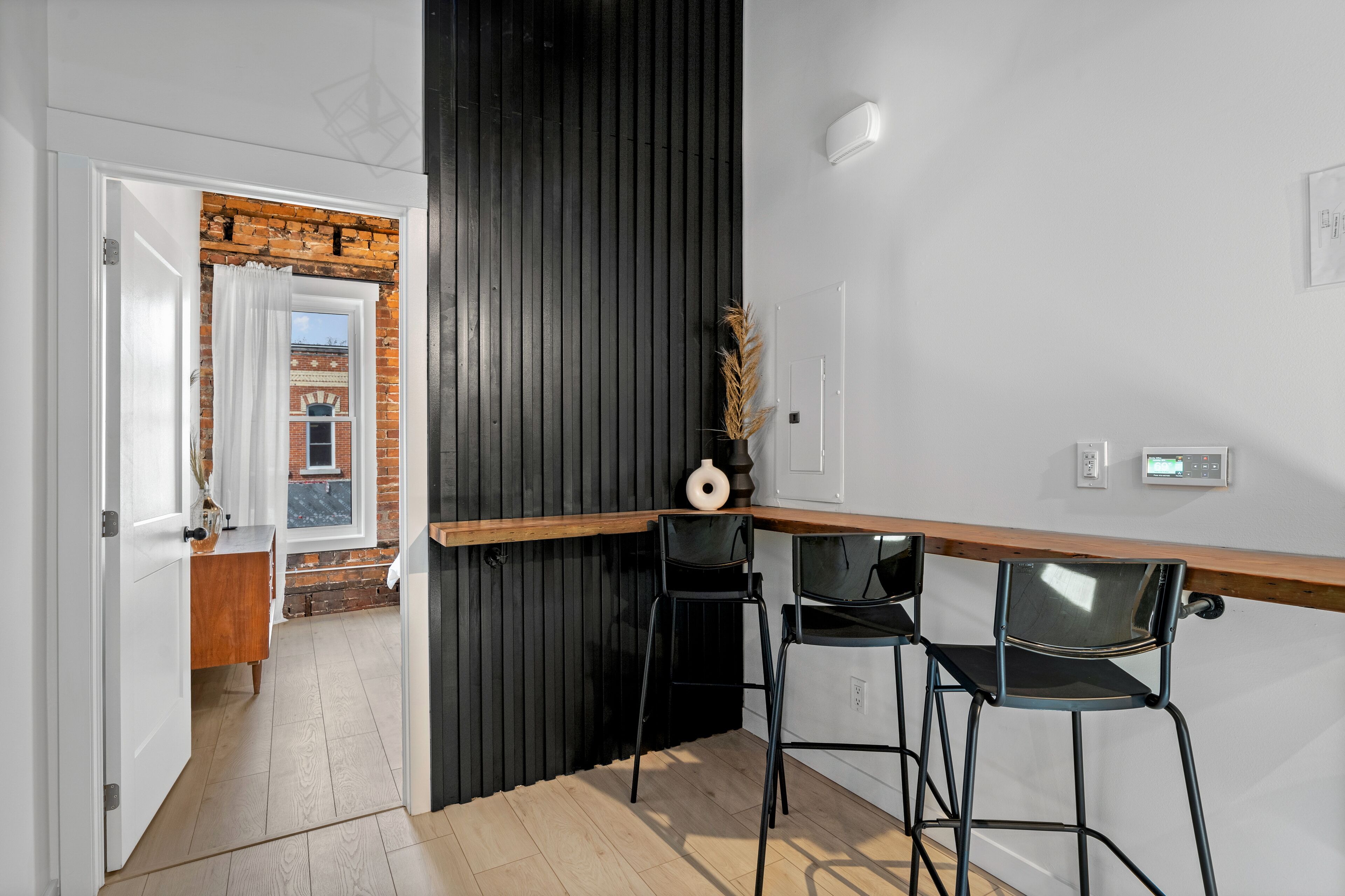 A modern kitchen with a wooden countertop, black bar stools, and a view of a brick building through an open door.