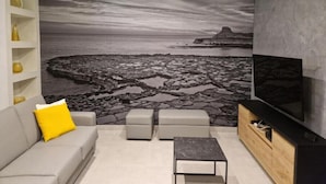 A living room with a large TV, grey sofa, ottomans, and a wall mural of a rocky coastline. - Red Rose.. (Iz-Zebbug)