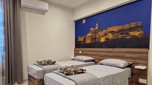 A hotel room with two beds, a large painting of a cityscape, and an air conditioning unit. - Red Rose.. (Iz-Zebbug)