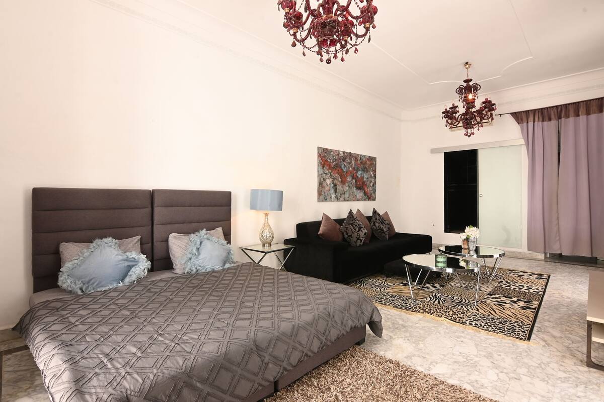 A modern bedroom with a large bed, a black sofa, a glass coffee table, and a chandelier.
