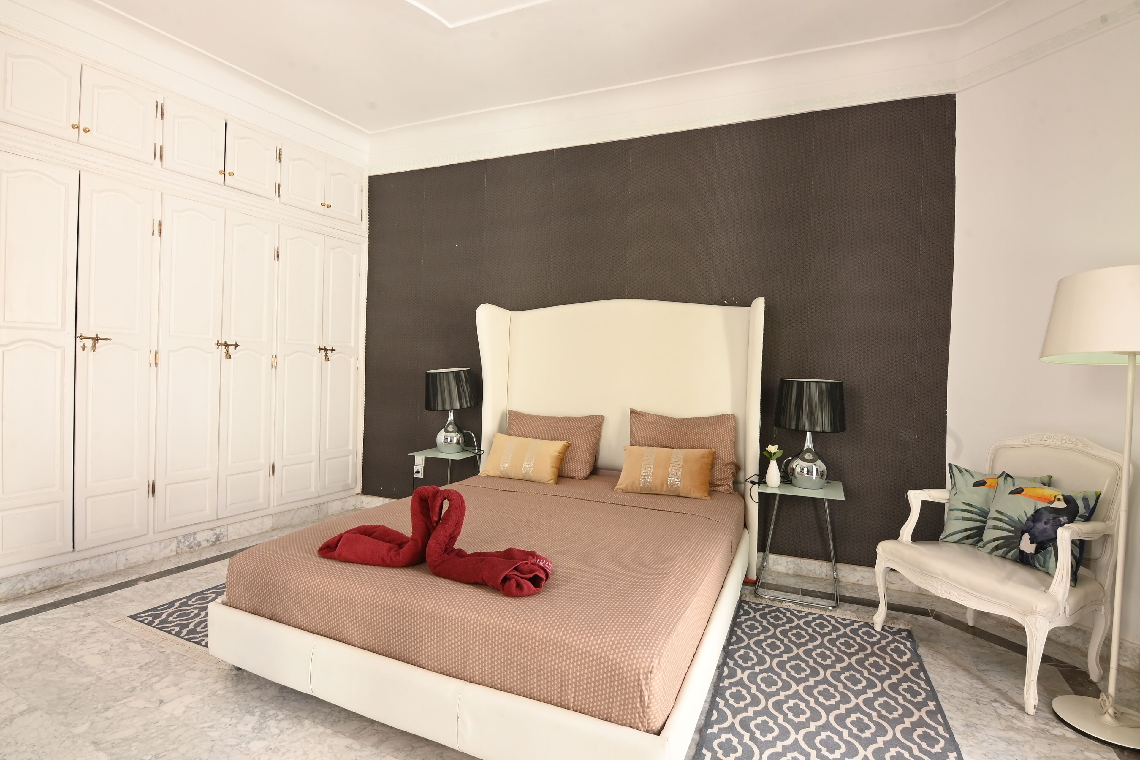 A hotel room with a large bed, a wardrobe, a nightstand with a lamp, a sofa, and a patterned rug.