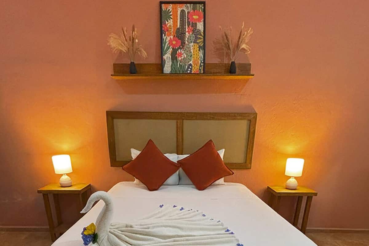 Una cama cuidadosamente tendida con una colcha blanca, dos almohadas rojas y una decoración ornamental con forma de cisne. Hay dos lámparas de noche, una obra de arte enmarcada sobre la cama y un cabecero de madera.