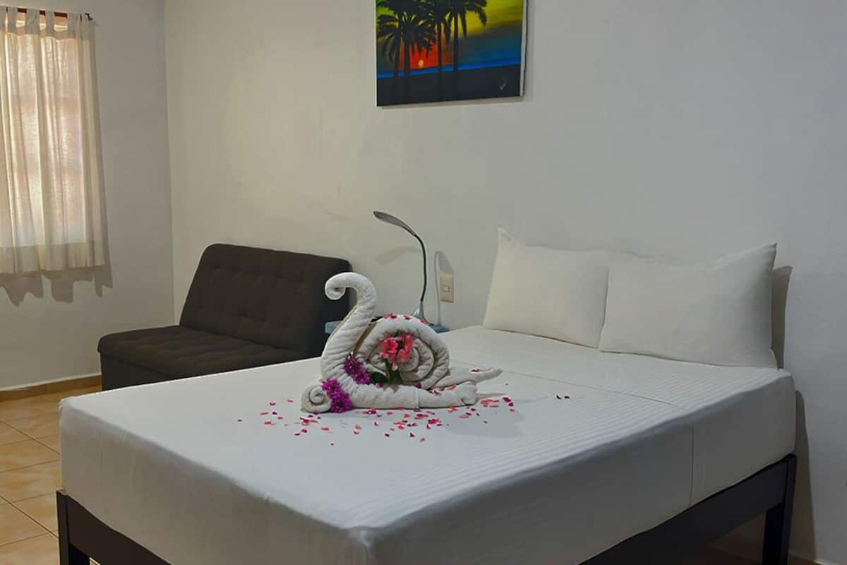 Una cama bien hecha con una toalla decorativa con forma de cisne, un cuadro de palmeras en la pared y un ventilador de techo.