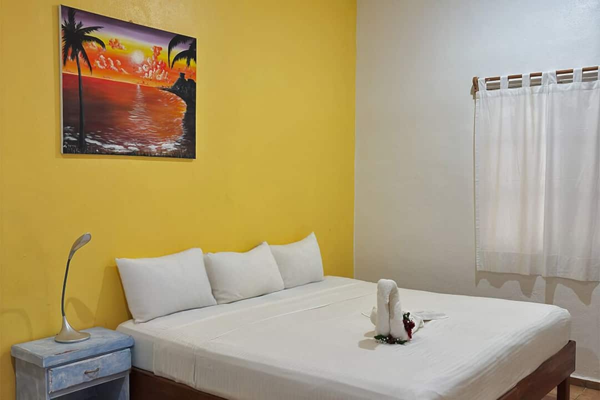 Un dormitorio con cama, almohadas blancas, una mesita de noche con lámpara, un cuadro de un atardecer y una cortina blanca.