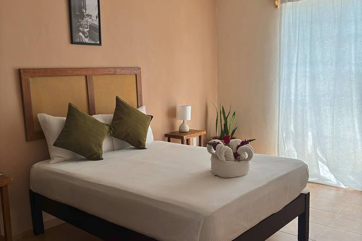 Una cama bien hecha con cabecera de madera, una lámpara de noche y una mesita con una planta.
