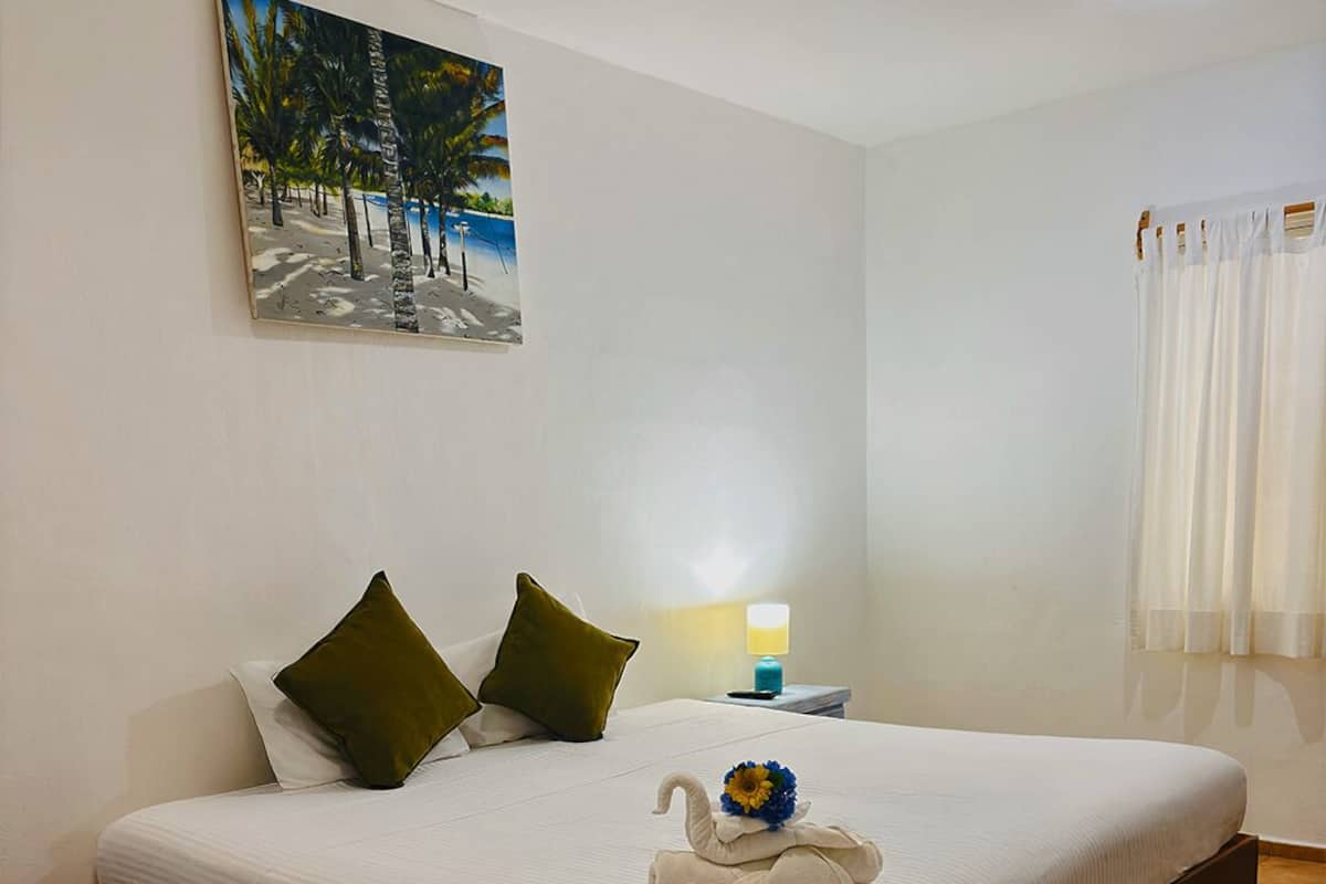 Habitación de hotel con una cama grande, dos almohadas verdes, una manta blanca, una mesita de noche con una lámpara y un cuadro de una playa.