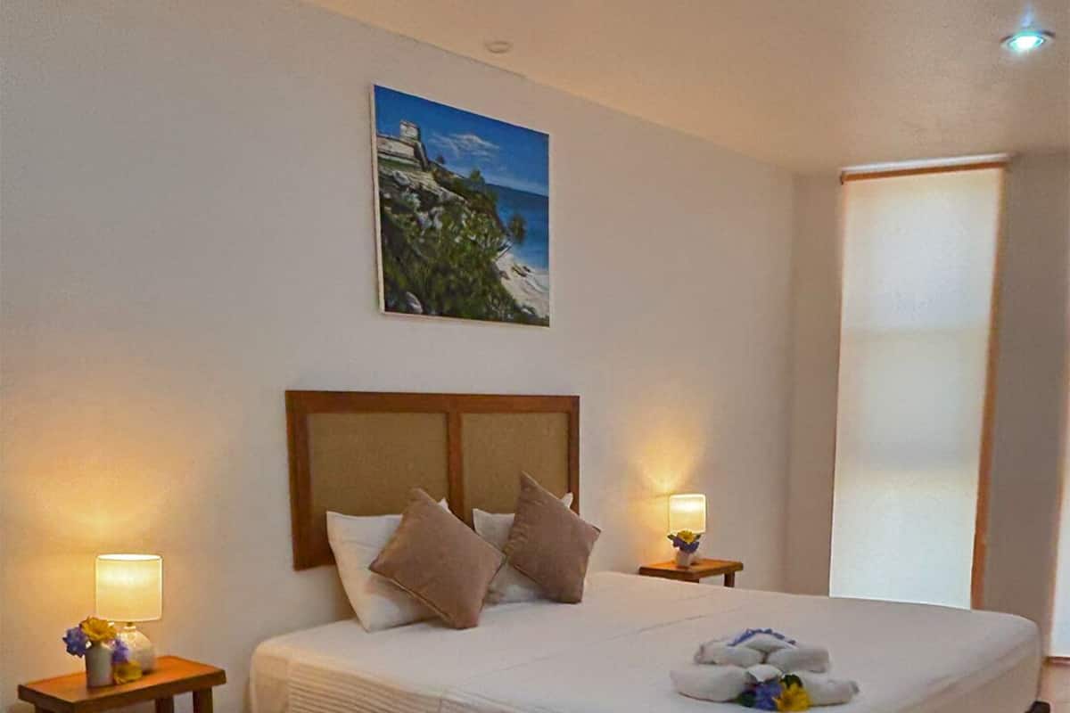 Un dormitorio ordenado con una cama grande, dos mesitas de noche con lámparas, un cuadro en la pared y un ventanal amplio.