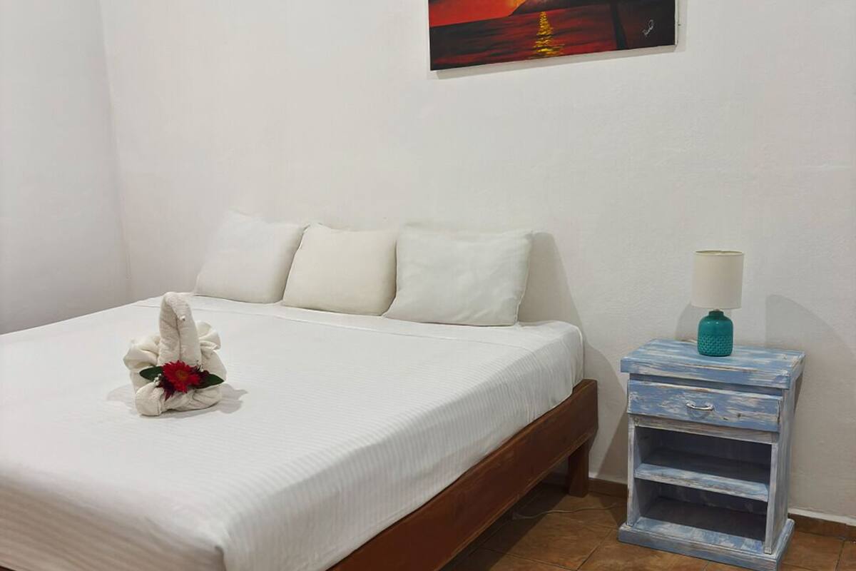 Una cama bien hecha con un cojín y un adorno en la cabecera, una mesita de noche con una lámpara, y un cuadro de un atardecer en la pared.