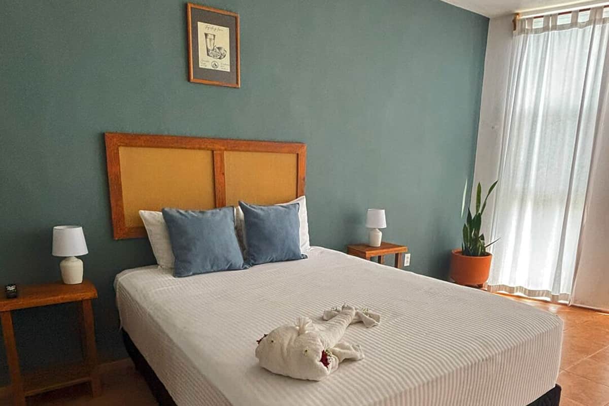 Una habitación de hotel con una cama grande, dos mesitas de noche con lámparas, un cuadro enmarcado en la pared y una planta en maceta.