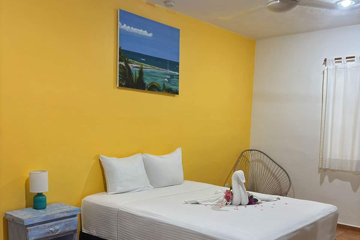 Un dormitorio con cama, mesita de noche, silla, ventilador de techo y un cuadro de una playa.