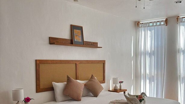 Habitación de hotel con una cama grande, ventilador de techo, cabecera de madera, mesita de noche con lámpara y ventana con cortinas.