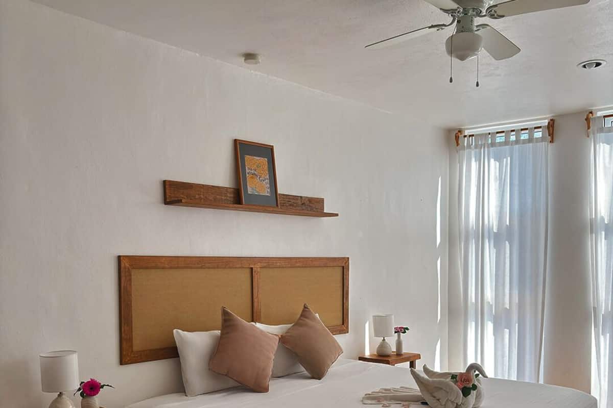 Habitación de hotel con una cama grande, ventilador de techo, cabecera de madera, mesita de noche con lámpara y ventana con cortinas.