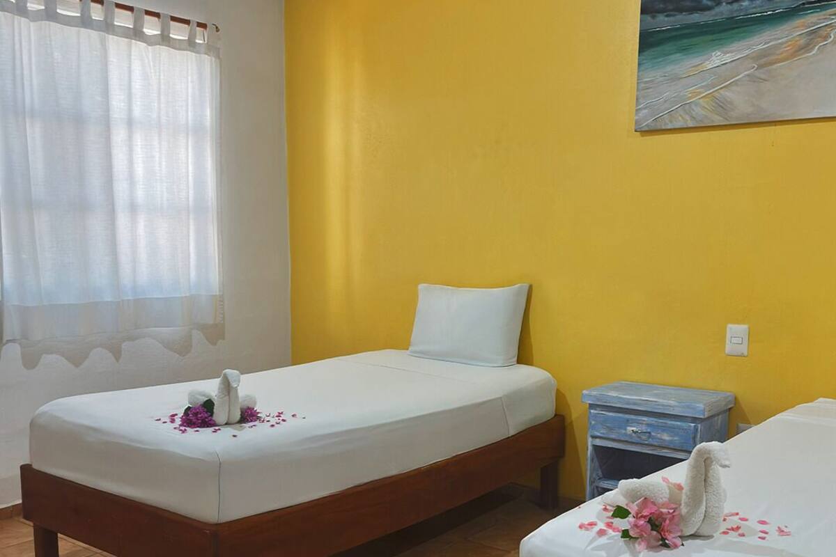 Habitación de hotel con dos camas individuales, un cuadro de una escena de playa y una ventana con cortinas translúcidas.
