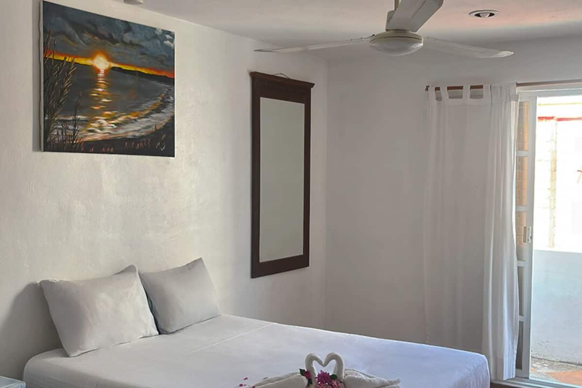 Habitación de hotel con cama, un cuadro en la pared, ventilador de techo y vista por la ventana.